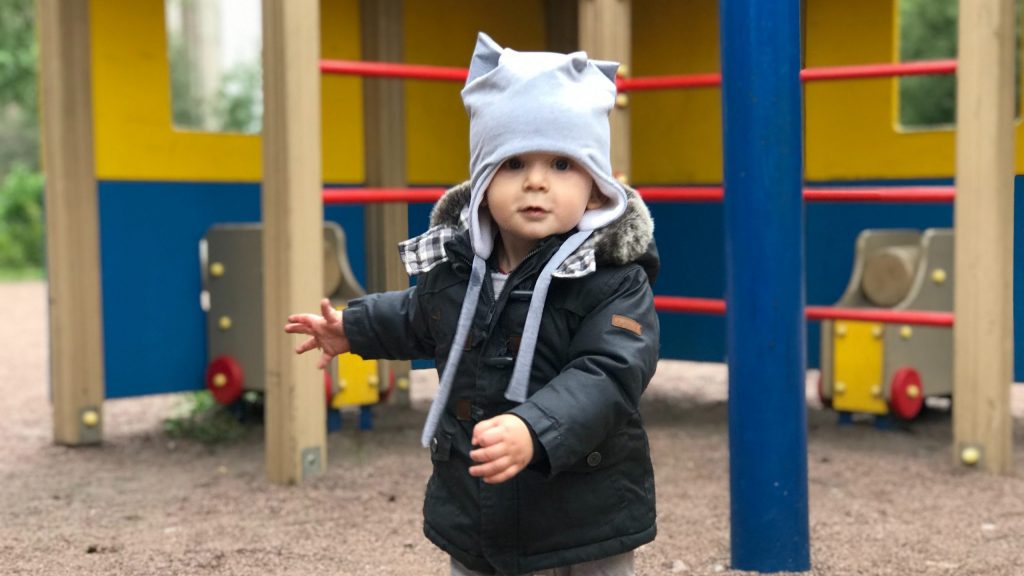 child in black jacket and white cap standing near yellow blue and red playground during daytime