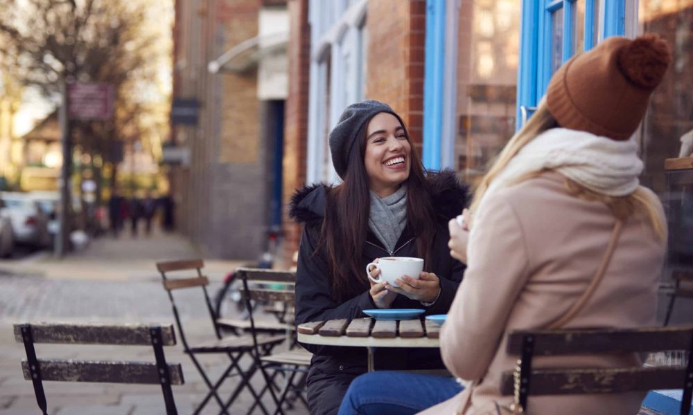 Two Female Friends Meeting Sitting Outside Coffee Shop On City High Street Two Female Friends Meeting Sitting Outside Coffee Shop On City High Street